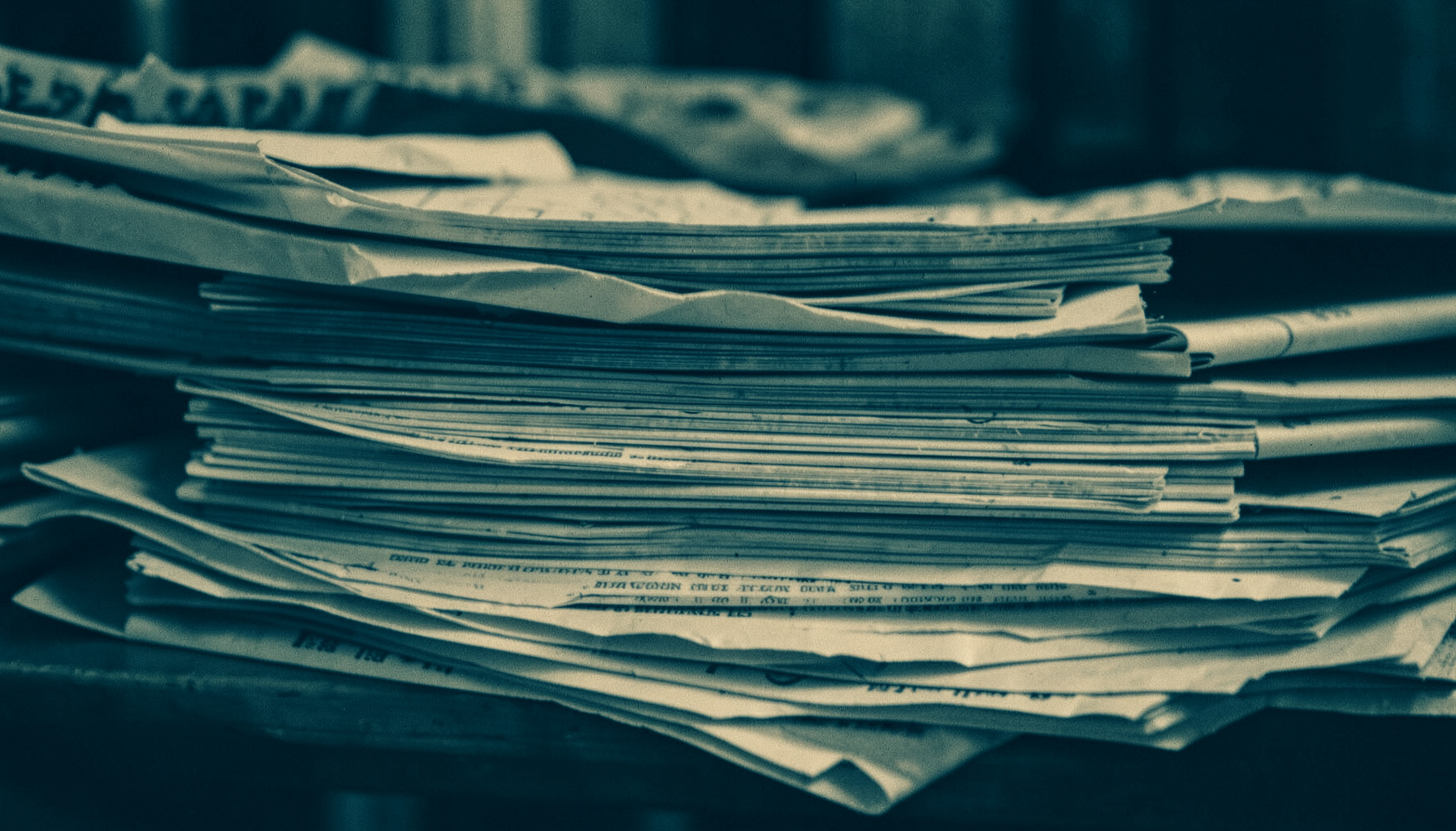 A moody duotone photograph of stacked newspapers in deep teal and warm cream tones, evoking a quiet newsroom.