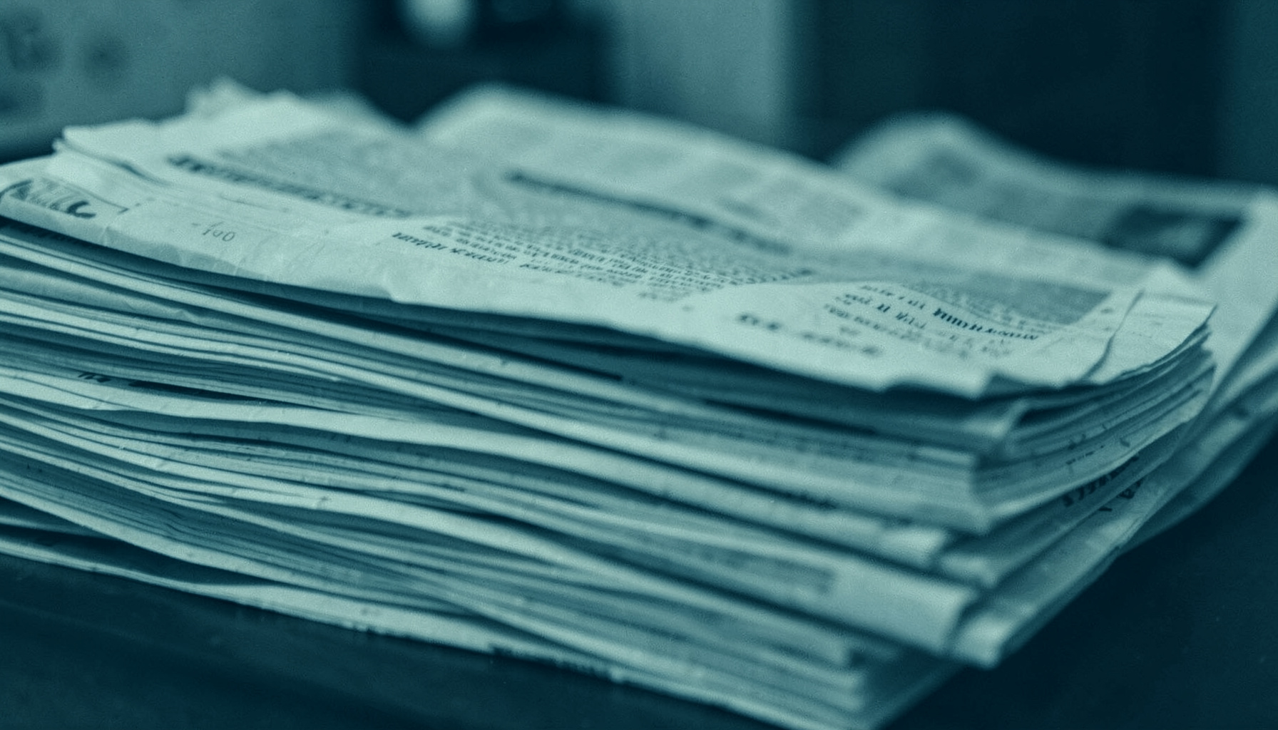 Duotone photograph of a stack of newspapers in muted teal and off-white, suggesting a quiet newsroom mood.